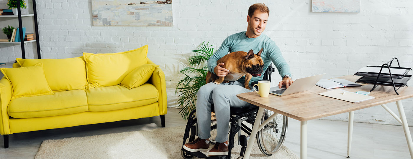 Man in a wheelchair works on his computer while his dog sits in his lap