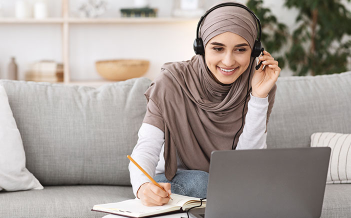 Smiling Muslim woman takes notes as she learns from her laptop