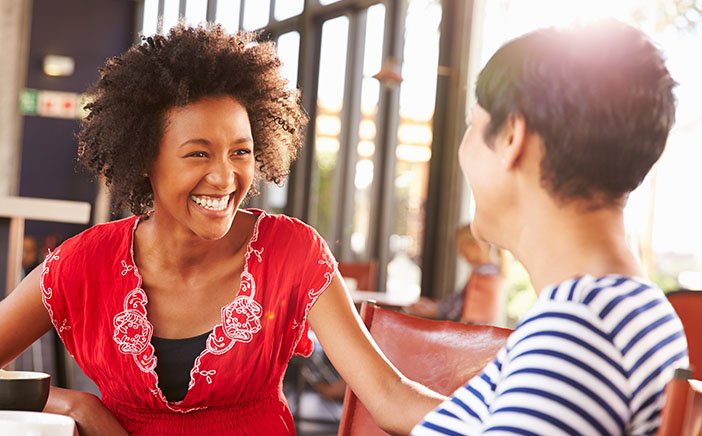 Two Black women enthusiastically engage in a conversation together while sitting down in a cafe