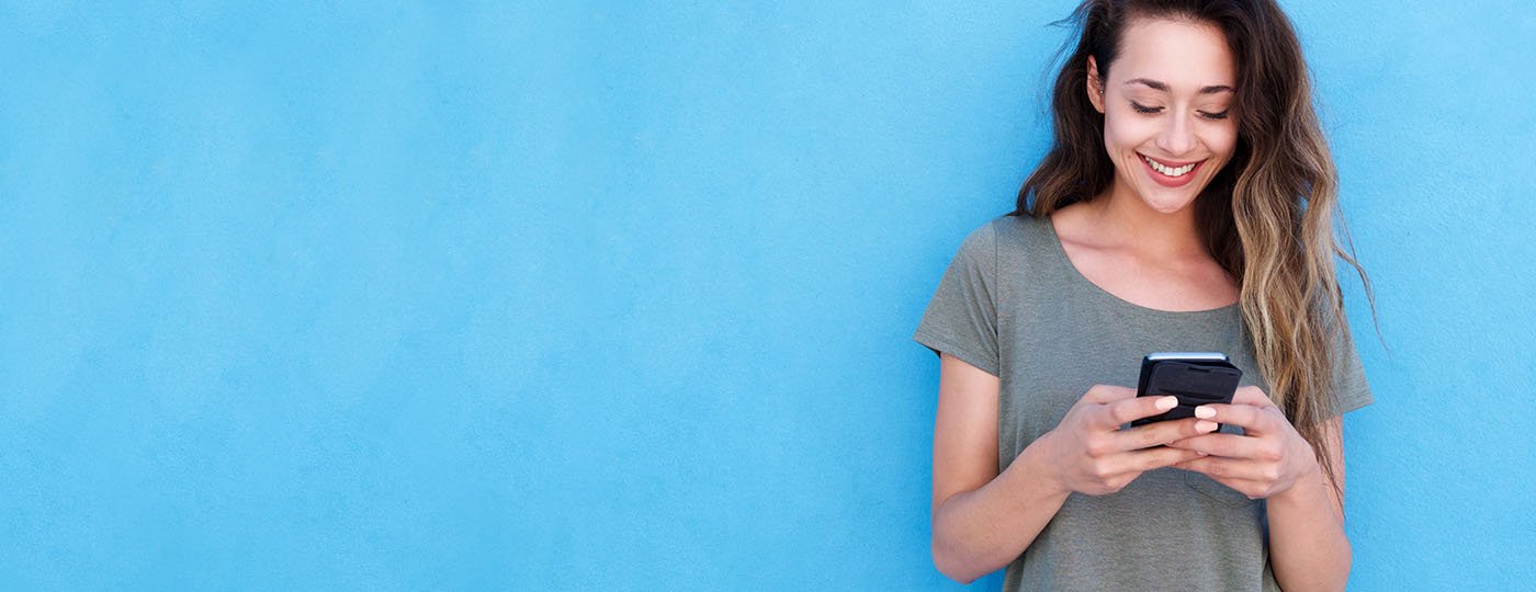 Young woman standing against a blue wall and smiling at her mobile phone in her hands