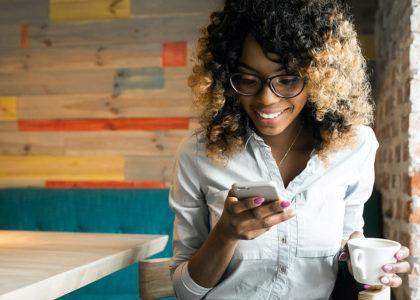 Black woman sits in a cafe and smiles at her smartphone in her hand while holding a cup of coffee