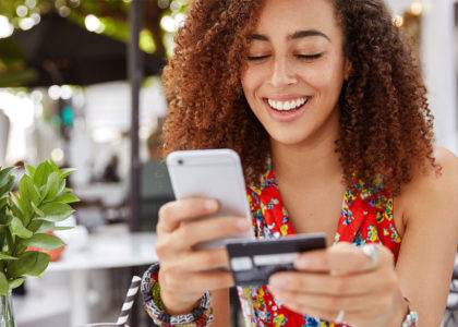 Young Black woman sits outside a cafe and smiles at her smartphone as she makes an online purchase