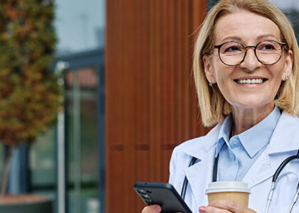 Portrait of a mature older female woman doctor or nurse and health care worker using a smartphone mobile phone in front of hospital or clinic outside