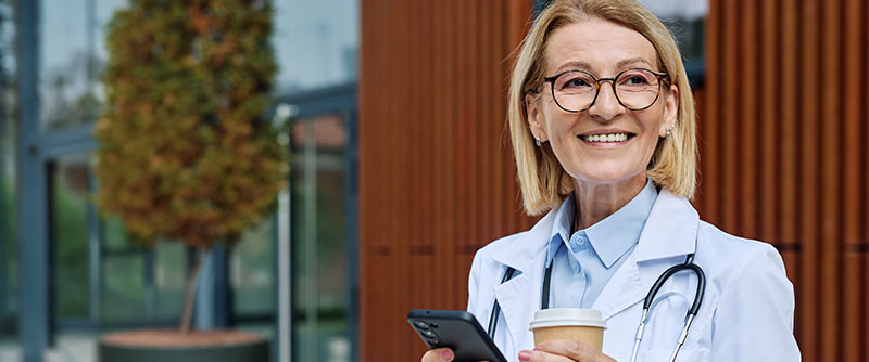 Portrait of a mature older female woman doctor or nurse and health care worker using a smartphone mobile phone in front of hospital or clinic outside