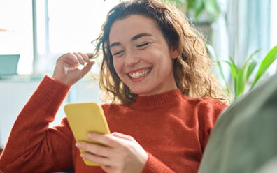 Young woman laughs at her phone as she sits on her couch at home