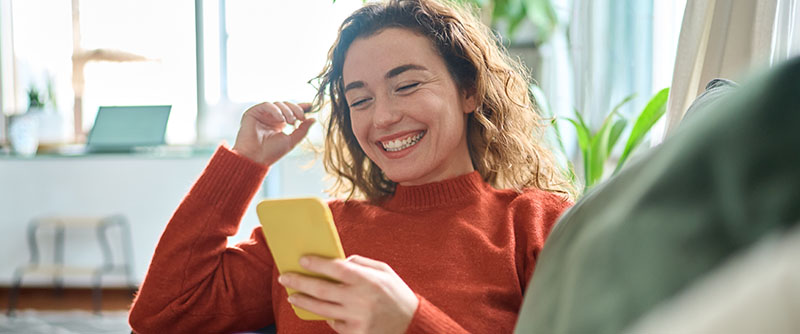 Young woman laughs at her phone as she sits on her couch at home