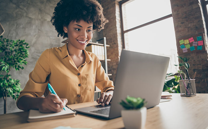 Black young woman smiling at her laptop as she takes notes on a notepad
